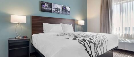 A hotel room with a bed, two bedside lamps, a nightstand, a wall art piece, and a window with curtains.