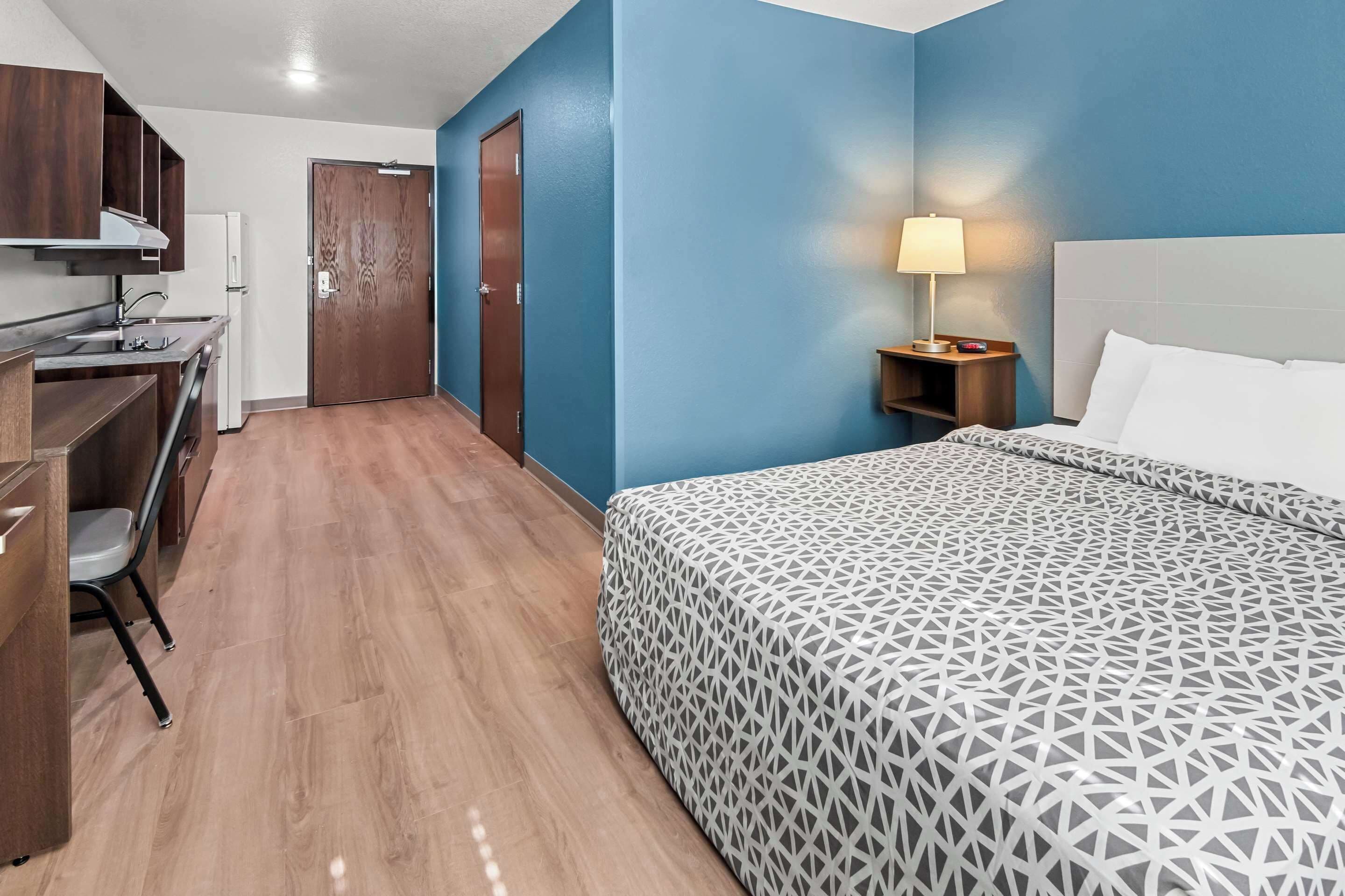A hotel room with a bed, a desk with a chair, a nightstand with a lamp, and a wardrobe.