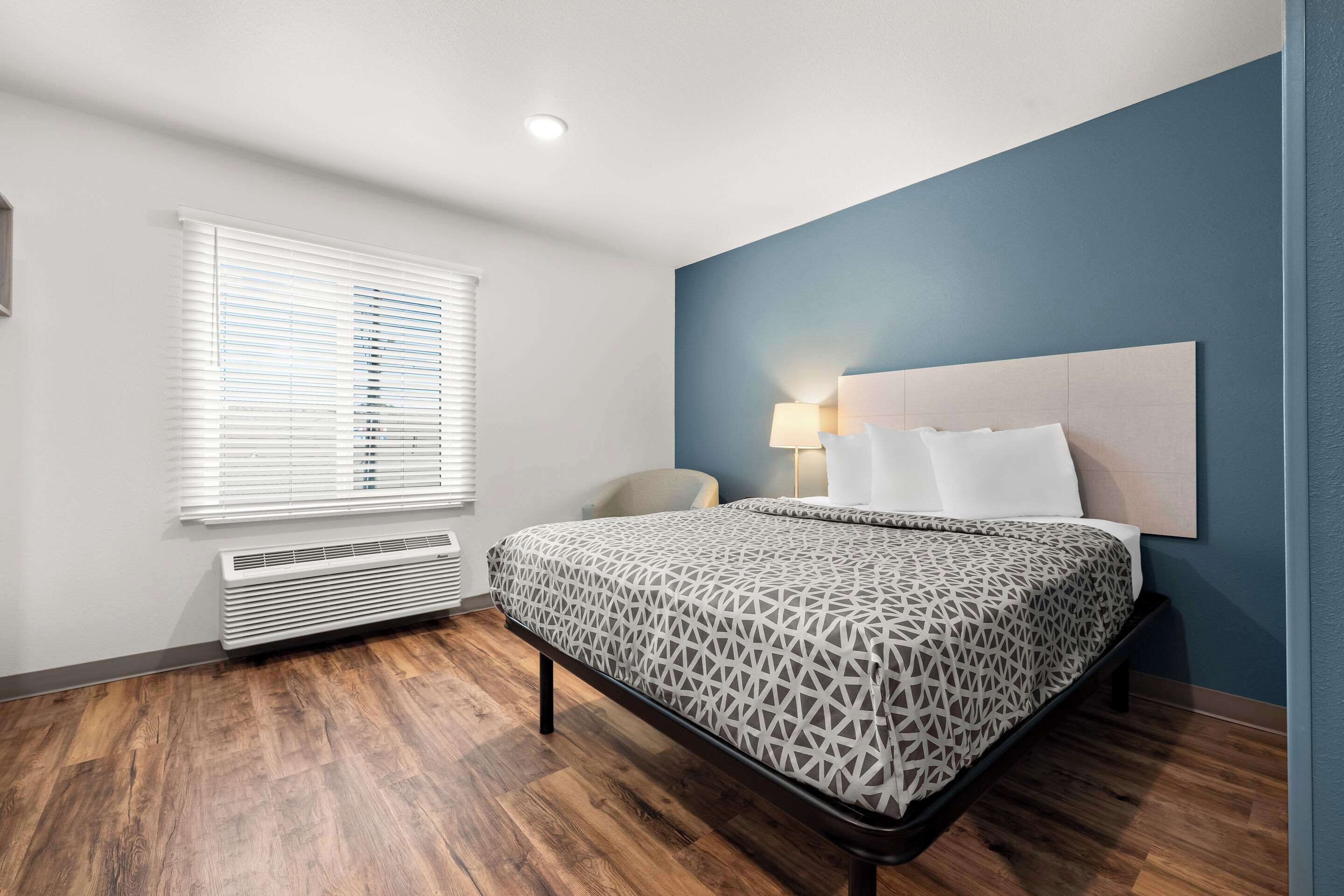 A modern hotel room with a large bed, a chair, a desk, and a window with blinds.