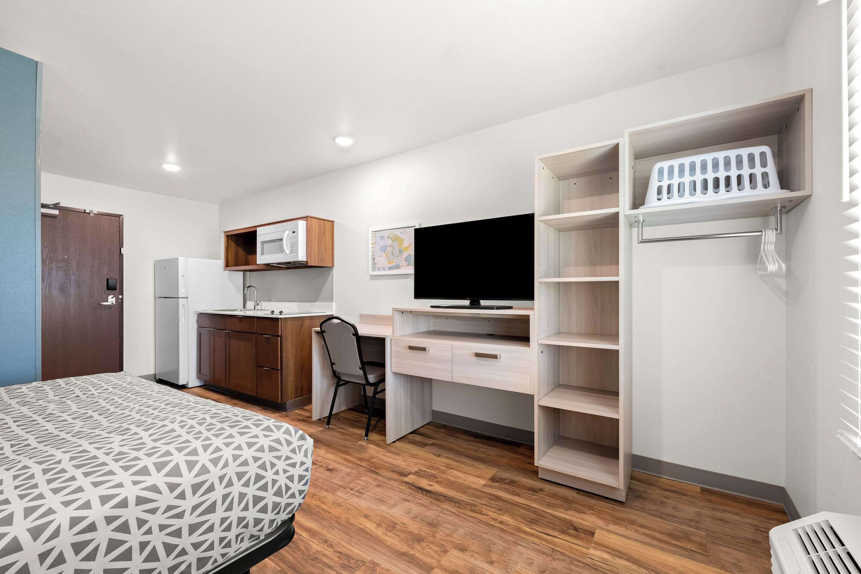A compact room with a bed, kitchenette, desk, and shelving unit.