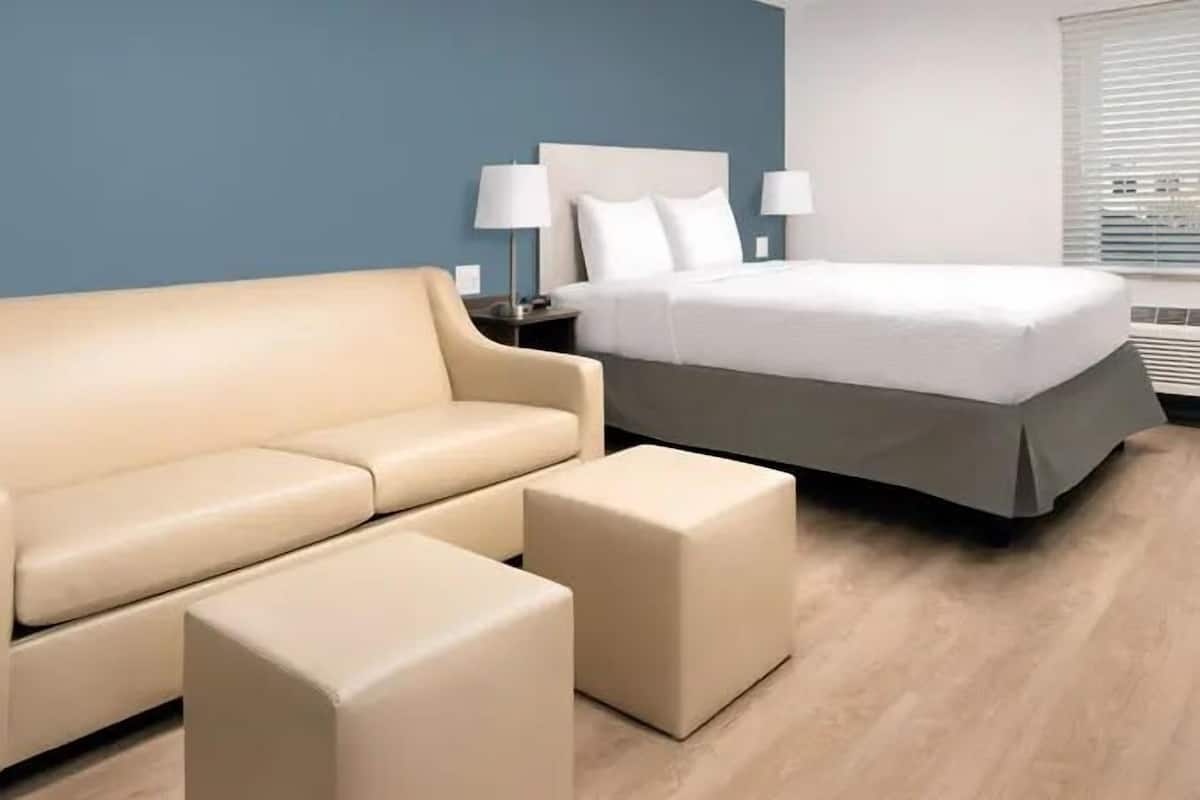 Een hotelkamer met een beige bank, een bijpassend poufje, een bed met wit beddengoed en twee bedlampjes.