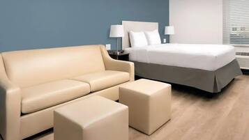 A hotel room with a beige sofa, a matching ottoman, a bed with white bedding, and two bedside lamps.