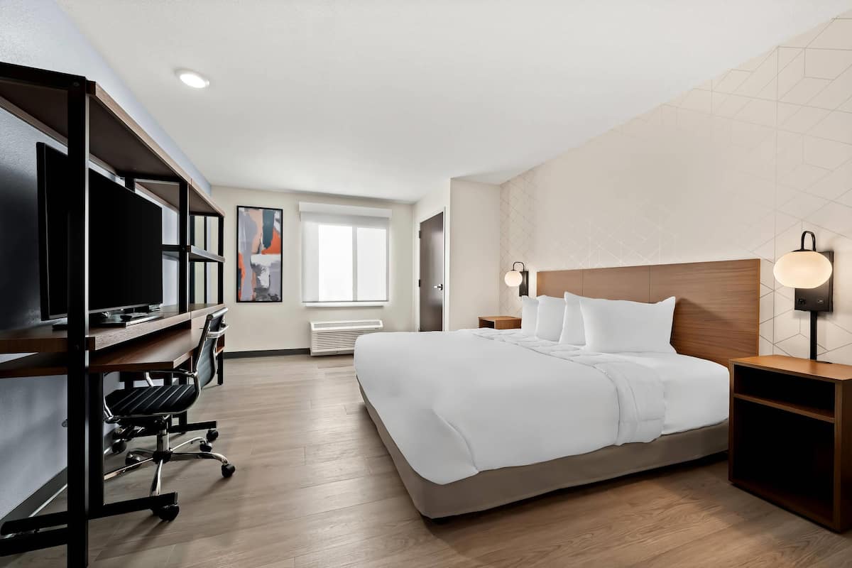 A modern hotel room with a large bed, a desk with a chair, a TV, and a bedside table with lamps.