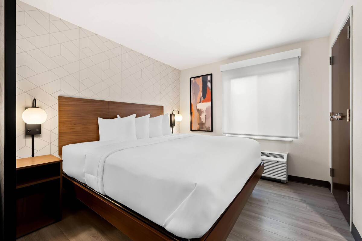 A hotel room with a large bed, wooden headboard, bedside lamps, a window with blinds, and a wall-mounted air vent.