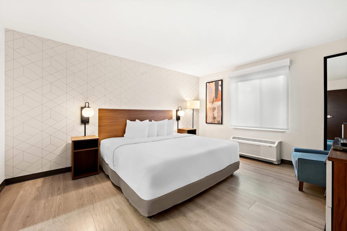 A modern hotel room with a large bed, wooden headboard, two bedside tables with lamps, a wall-mounted air conditioner, and a large window with blinds.