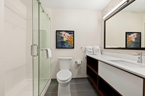 Un baño moderno con cabina de ducha de vidrio, inodoro blanco, lavamanos con espejo y un cuadro abstracto enmarcado en la pared.