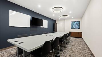 Un salón de conferencias con una larga mesa, sillas negras, una pizarra y un televisor montado.