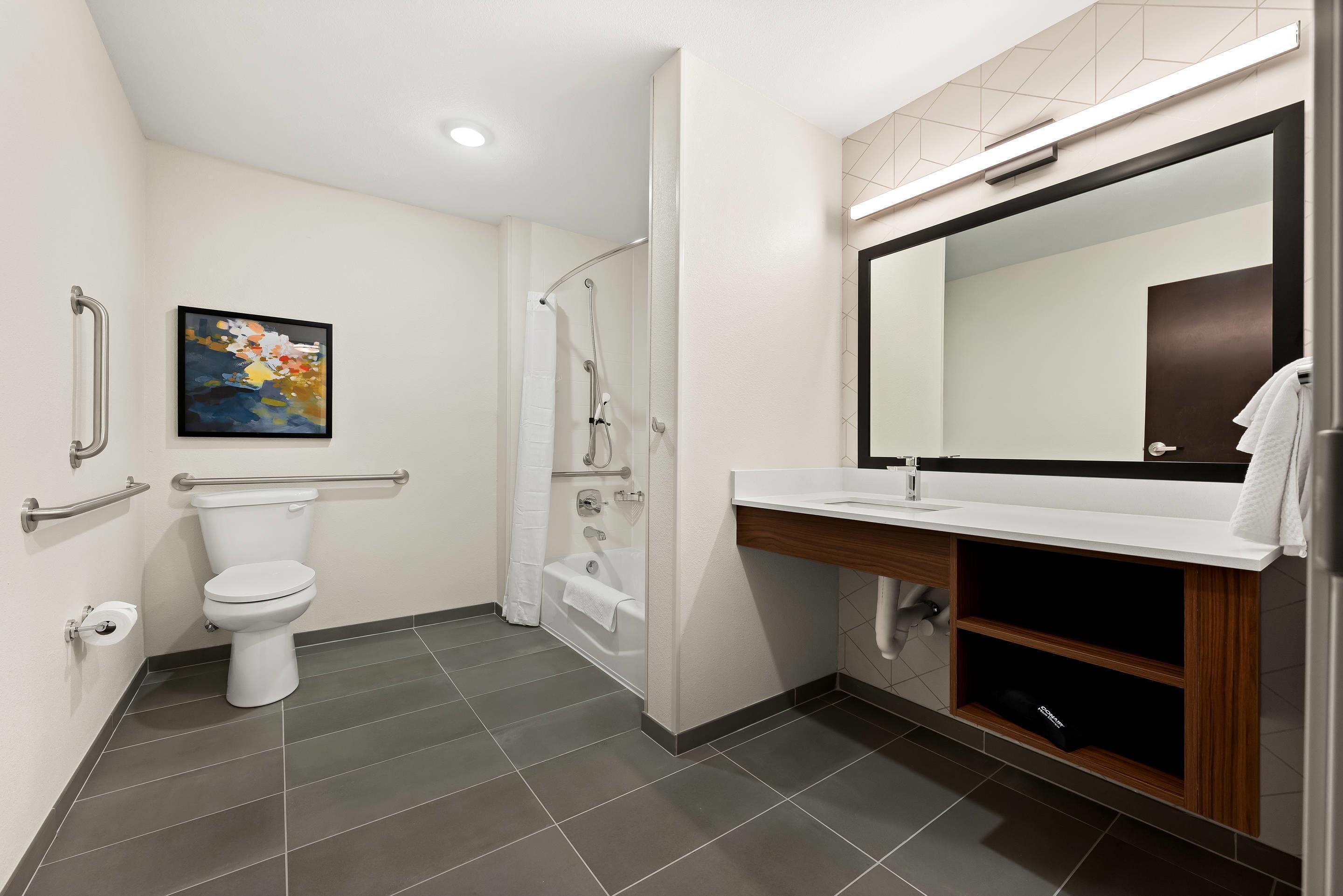 Un baño moderno con inodoro, ducha y lavamanos, equipado con barras de apoyo y un espejo grande.