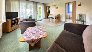 Un salon comprenant un canapé, une petite table recouverte d’une nappe à carreaux, un téléviseur posé sur un meuble en bois, et un coin repas avec une table et des chaises.