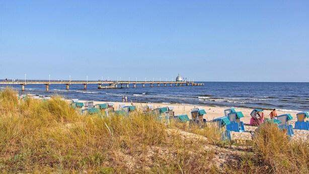 Une plage bordée de rangées de chaises de plage bleues et d’une jetée qui s’avance dans la mer.
