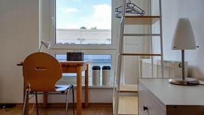 Une petite pièce bien rangée, avec un bureau en bois, une chaise, un radiateur, une lampe et une fenêtre donnant sur des immeubles.