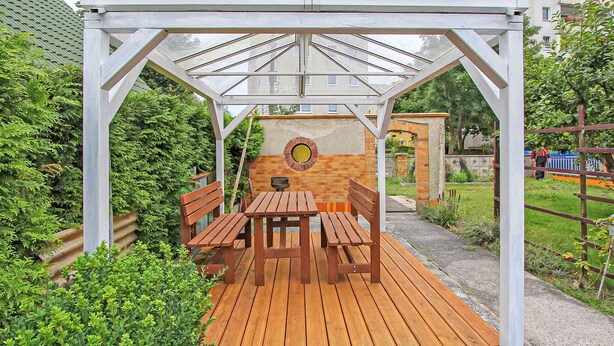 Eine Holzterrasse mit einer Pergola, Bänken und einer Backsteinmauer mit rundem Fenster.
