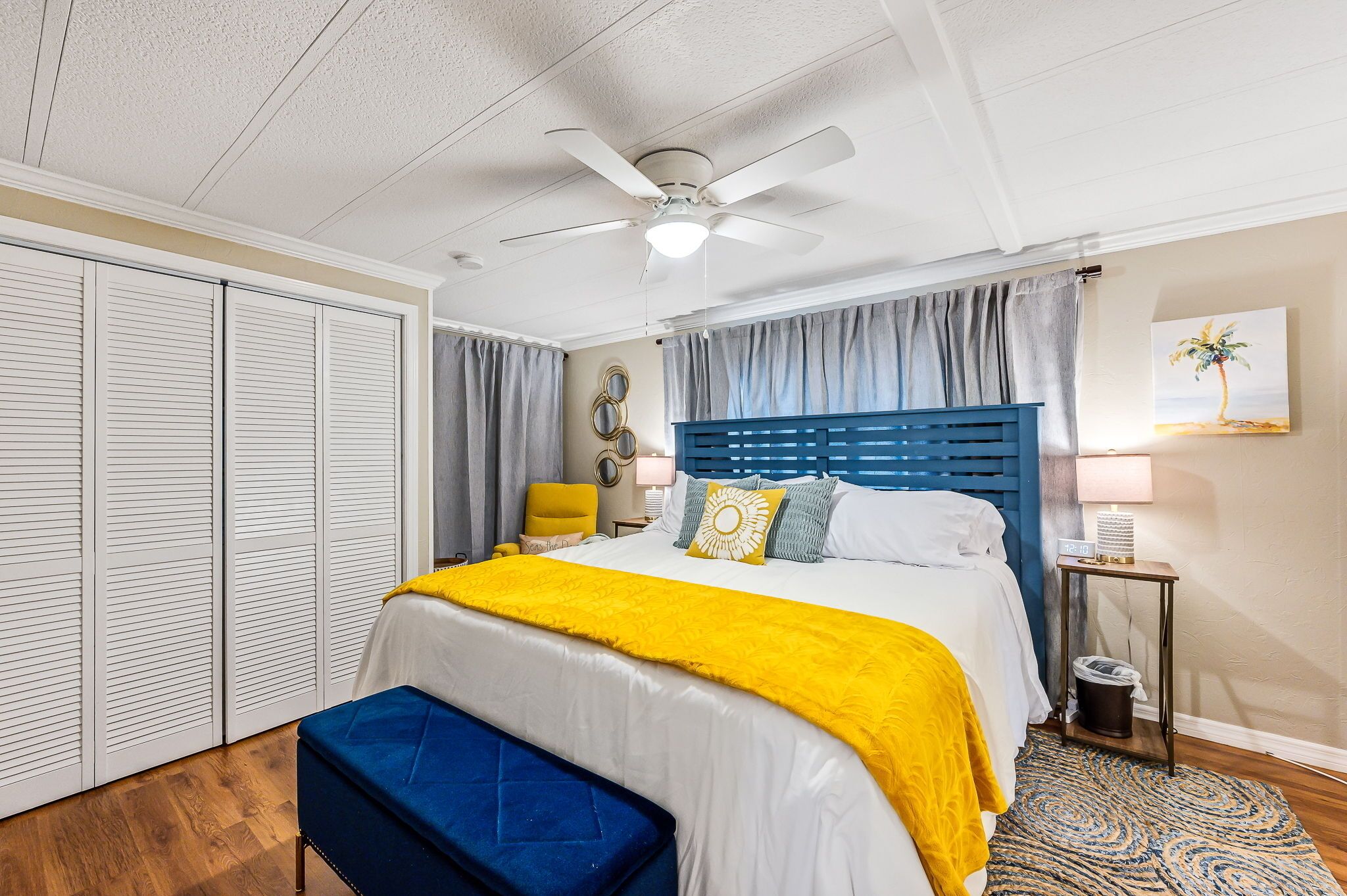 A bedroom with a bed, a ceiling fan, a bedside table, a lamp, a trash can, a bench, and a window with curtains.