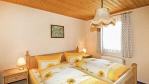 Een slaapkamer met een houten plafond, een bed met bloemenbeddels, een nachtkastje met een lamp, een raam met gordijnen en een foto aan de muur.