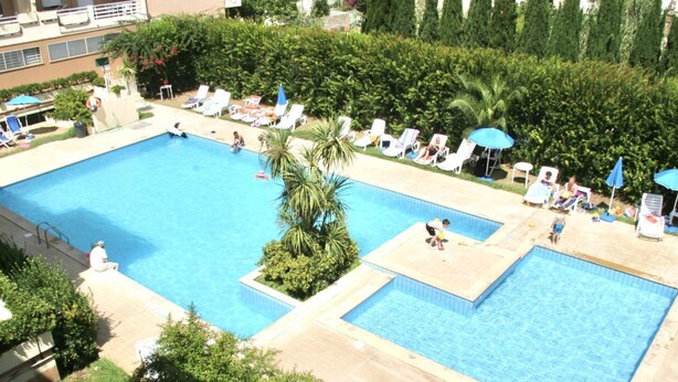 Une grande piscine extérieure divisée en plusieurs zones, entourée de chaises longues et de parasols, et jouxtant un bâtiment doté de balcons.