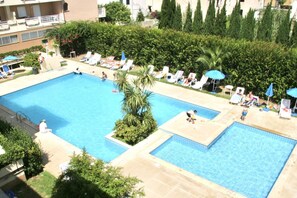 A large outdoor swimming pool with multiple sections, surrounded by lounge chairs and umbrellas, and adjacent to a building with balconies.