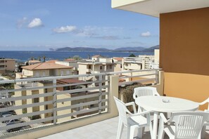 A balcony with a white table and chairs, overlooking a coastal view with buildings and mountains in the distance.