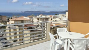 Un balcon avec une table et des chaises blanches, offrant une vue sur la côte, avec des bâtiments et des montagnes visibles au loin.