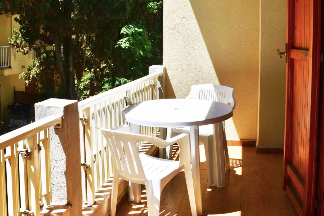 Een balkon met een witte plastic tafel en stoelen, een houten hekwerk en een uitzicht op bomen en een gebouw op de achtergrond.