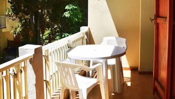 Een balkon met een witte plastic tafel en stoelen, een houten hekwerk en een uitzicht op bomen en een gebouw op de achtergrond.