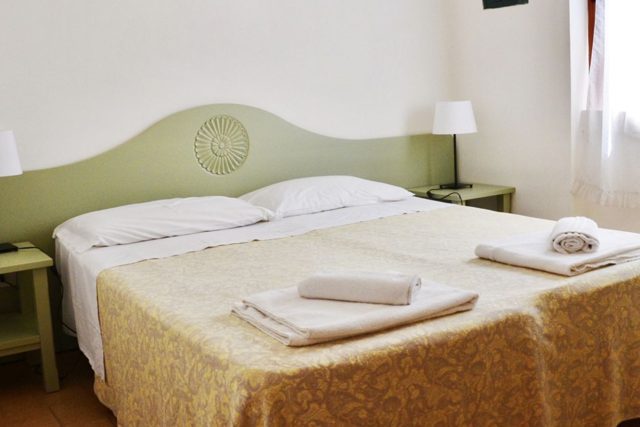A neatly made bed with white linens, a green headboard, and a patterned bedspread. There are two bedside tables with lamps, and a rolled towel on the bed.