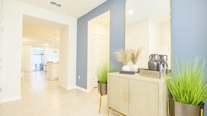 A modern hallway with a light-colored floor, a blue accent wall, and a large mirror reflecting the room. There is a decorative cabinet with gold legs and a potted plant beside it.