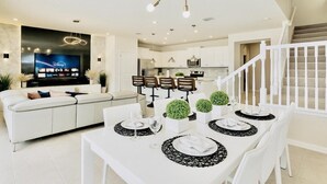 A modern living room with a flat-screen TV, a dining area with a white table set for four, and a staircase leading to an upper level.