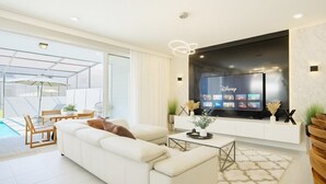 A modern living room with a white sofa, a coffee table, and a flat-screen TV mounted on the wall. There is a pool visible through the glass doors.