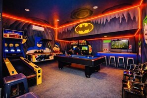 Quarto com mesa de bilhar, máquinas de fliperama e um grande logotipo do Batman na parede.