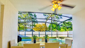 A covered patio with a glass table and wicker chairs, overlooking a swimming pool and a fenced yard. - Fantastic 5BD House w/Private Pool - 9mi/Disney! (Clermont)