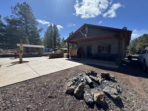 Situated in a natural setting, this rustic lodge features a sloped roof and chimney, with a wooden barrel and benches nearby.