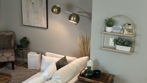 A modern living room with a sofa, a side table, a wall-mounted shelf with plants, a framed picture, and a lamp.