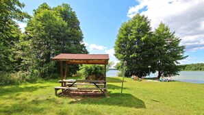 Un espace de pique-nique doté d’un abri en bois, d’une table de pique-nique et de bancs, le tout entouré d’arbres et bordé par un lac.