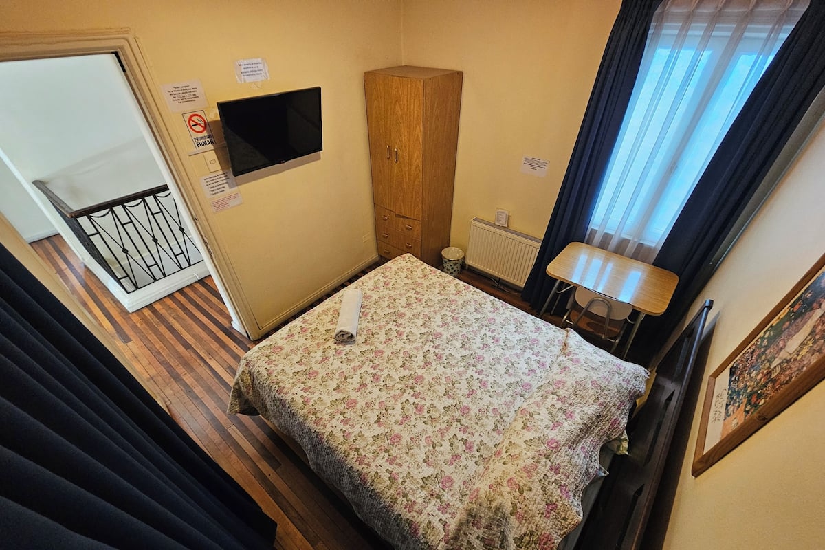 Un dormitorio pequeño y ordenado con cama, armario de madera, televisor, radiador y ventana con cortinas.