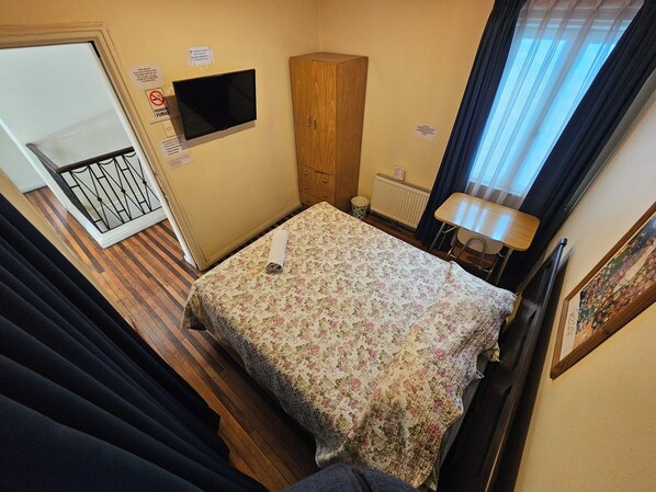 A small, neatly arranged bedroom with a bed, a wooden wardrobe, a television, a radiator, and a window with curtains.