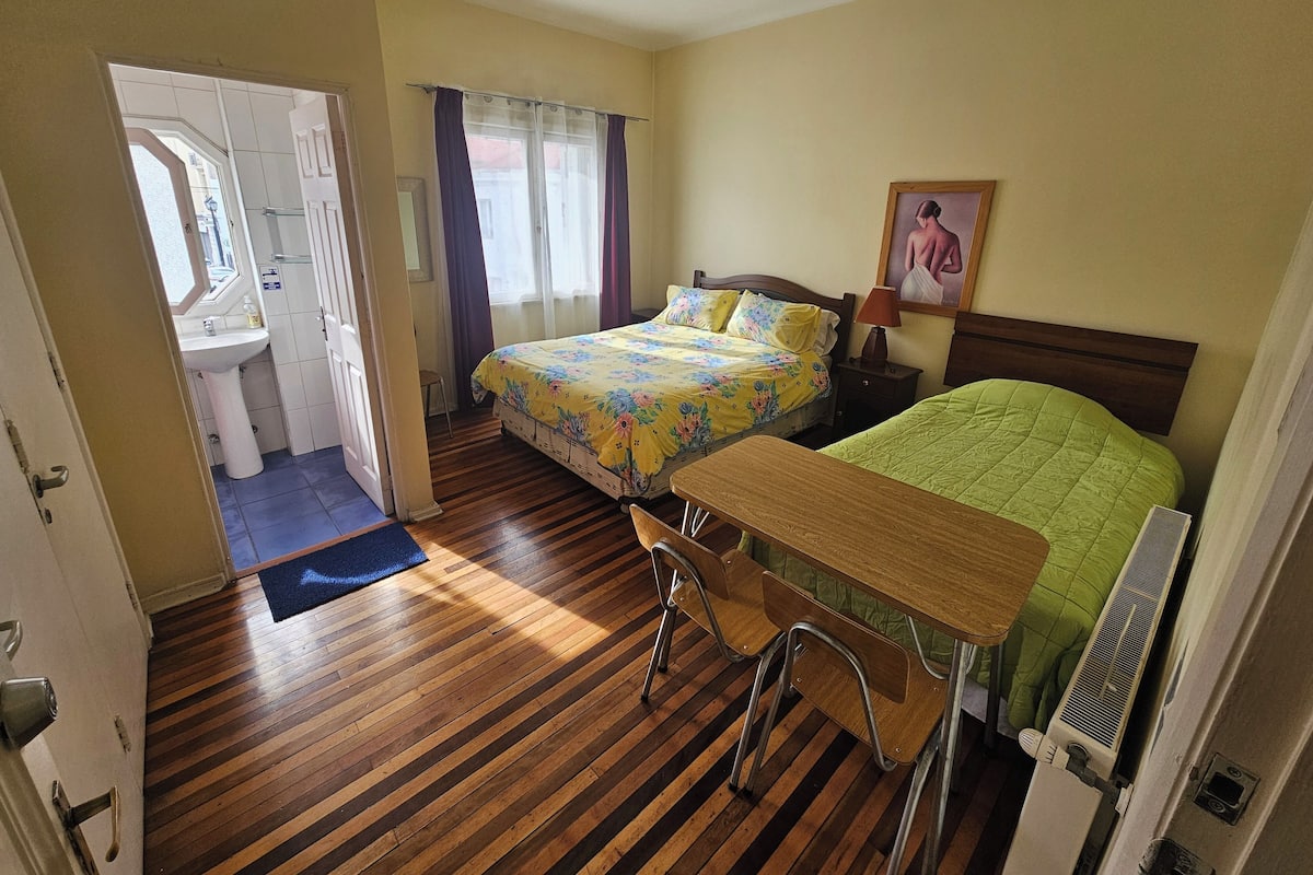 Un dormitorio con dos camas, un escritorio y una silla. Hay una ventana con cortinas, un radiador y un cuadro en la pared.