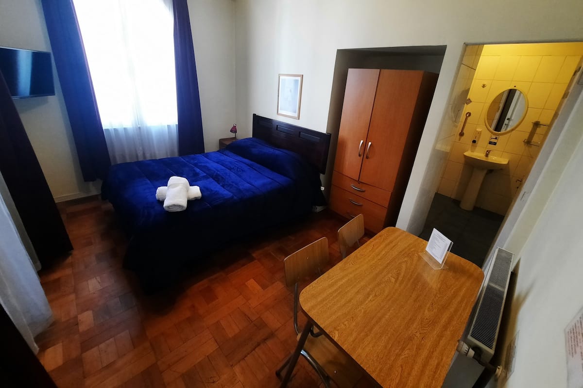 Habitación de hotel pequeña con cama, una mesa de madera, una silla, armario, lavamanos y espejo.