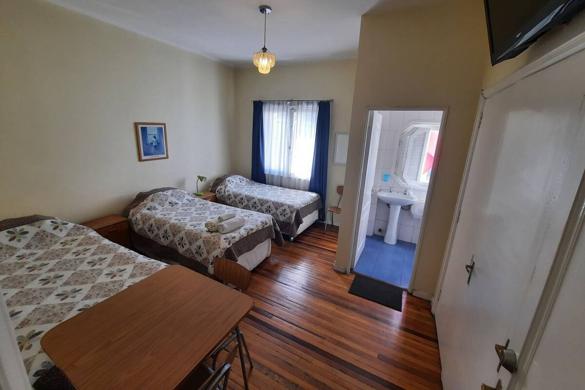Habitación de hotel con dos camas, una mesa de madera, un escritorio, una lámpara de escritorio, un aplique y un baño visible a través de una puerta abierta.
