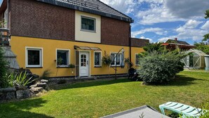 A two-story house with a yellow facade, a brick section, and a small front yard with a lawn, a stone wall, and a green bush. - Gorgeous apartment in Lychen with sauna (Lychen)