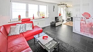 Een moderne woonkamer met een rode bank, een salontafel en een televisie. Er zijn grote ramen met gordijnen, een eethoek met een tafel en stoelen, en een keuken met een koelkast en kastjes.