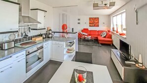 Een moderne keuken met witte kasten, een ingebouwde oven en een koffiezetapparaat. Er is een zithoek met een rode bank en een televisie.