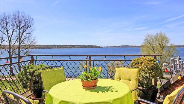 Een terras met een tafel bedekt met een groene tafelkleed, omringd door stoelen en een bloempot. Een gaashekwerk scheidt het terras van het water.