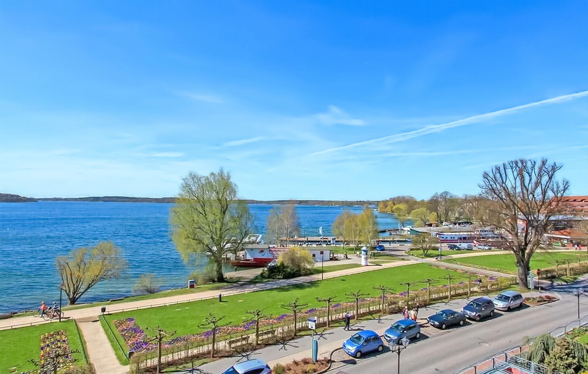 Vista del lago con zona de estacionamiento, camino peatonal y área verde con árboles.