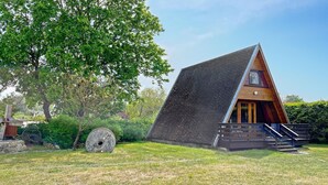Una moderna baita a tetto triangolare con esterno in legno, immersa in un prato con un albero grande e un mulino a pietra nelle vicinanze.