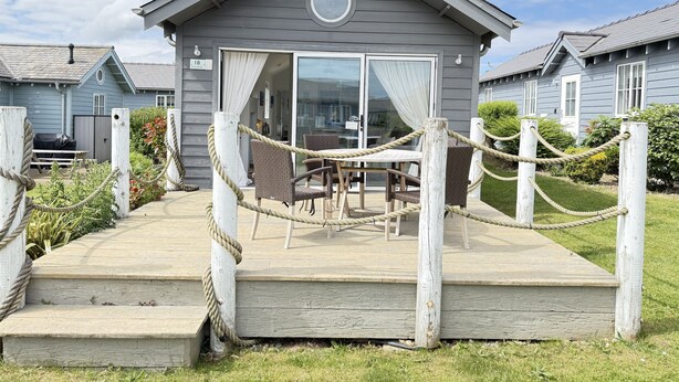 A wooden deck with a rope barrier leading to a building with a glass door and a circular window. - Sand Dollar (new for 2024) -  a beach house that sleeps 5 guests  in 2 bedrooms (Filey)