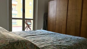 Quarto com uma cama, guarda-roupa de madeira e janela com vista para edifícios.