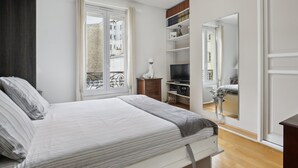 A bedroom with a bed, a TV, a bookshelf, and a window with curtains. - Studio St Dominique - Invalides - B'Your Home (Paris)