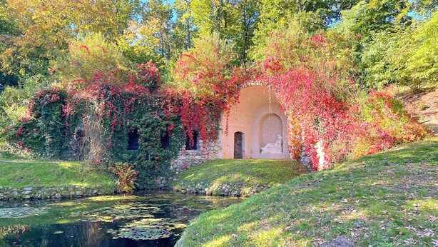 A serene garden with a pond, a stone wall, and a small arched structure surrounded by lush greenery and autumn foliage. - Ferienwohnung Rheinsberg (Rheinsberg)