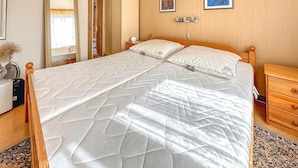 Een net opgemaakt bed met een gewatteerde matras, een houten hoofdbord en twee kussens. Een houten nachtkastje met een lamp staat ernaast.