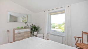 Et soveværelse med seng, kommode, spejl, radiator, et vindue med gardiner, en stol og en potteplante.